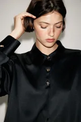 Camisa de satén color blanco, de corte clásico con cuello solapa, manga larga con puños y cierre frontal con botones dorados.