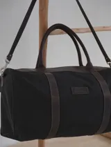 Bolso de viaje tipo duffel de lona negra con detalles en cuero tostado, asas de mano y correa ajustable para el hombro.