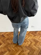 Pantalón de jean estilo wide leg con diseño de cintura cruzada y tiro bajo. Presenta un lavado azul medio con efecto desgastado y un detalle de rotura en la rodilla derecha.
