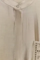 Camisa de cuello mao color beige, confeccionada en tejido satinado con brillo sutil, manga larga con puños anchos y un bolsillo de parche en el pecho con detalles bordados tonales. Cierre frontal con botones ocultos bajo solapa.
