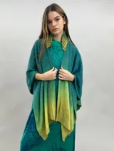Ruana de lana merino con diseño en degradé de tonos verdes. Presenta un cuello plisado artesanalmente en fieltro de lana merino, ofreciendo una textura suave y un acabado elegante.
