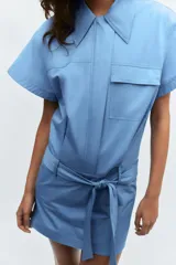 Vestido corto de estilo camisero color azul, con cuello solapa, manga corta y cierre frontal con botones. Presenta un bolsillo de plastrón con solapa en el pecho y un cinturón ajustable del mismo tejido en la cintura.