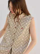 Chaleco clásico de corte cuadrado, color beige con estampado geométrico de círculos entrelazados y pespuntes en rojo. Cuenta con cuello camisero, cierre frontal de botones y un pequeño bordado en el pecho.