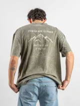 Remera de corte oversize color verde con efecto lavado, manga corta y cuello redondo. Presenta un estampado gráfico en la espalda con el diseño de una montaña y la frase 'Fresh air is free, don't be afraid of the high'.
