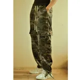 Pantalón cargo holgado con estampado camuflado en tonos verde militar, marrón y negro. Presenta cintura elástica ajustable con cordón blanco y bolsillos cargo laterales con cierre de botón a presión. Los tobillos son ajustables con cordones.