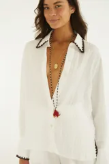 Camisa blanca de manga larga y calce relajado, confeccionada en tejido liviano de textura arrugada. Presenta cuello camisero, botonera frontal y detalles de ribete en zigzag marrón oscuro en cuello, puños y dobladillo.