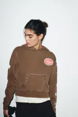 Sudadera marrón con capucha ajustable con cordones, manga larga con puños y bajo elástico. Presenta un estampado ovalado en el pecho con el texto "Texas USA" en rosa y azul, y un diseño de estrellas blancas en las mangas. El bolsillo canguro frontal tiene detalles de tachas y bordes deshilachados.