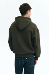 Campera con capucha y cierre frontal, color verde militar oscuro, de tejido pesado con interior cepillado suave. Presenta hombros caídos, cordón ajustable en la capucha y bolsillos canguro delanteros.