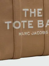 Cartera tipo tote bag Marc Jacobs modelo The Large Tote, color marrón con el texto "THE TOTE BAG MARC JACOBS" impreso en blanco en el frente. Confeccionada en lona resistente, cuenta con asas superiores dobles para llevar al hombro o en la mano.