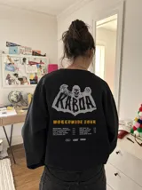 Buzo gris oscuro con efecto acid wash, corte regular fit y cuello redondo. Presenta una estampa grande en la espalda con diseño de calavera, el texto "Kaboa" y detalles de fechas de tour en tonos claros.