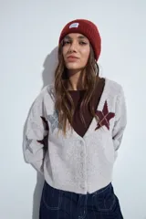 Cardigan corto tejido en color gris, con escote en V y cierre frontal de botones. Presenta un diseño estampado con estrellas y líneas geométricas en tonos oscuros y rojizos, con mangas largas y puños acanalados.