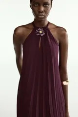 Vestido midi plisado color bordó, con cuello halter, broche de flor en el delantero y anudado en la espalda. Cuenta con forro interior.