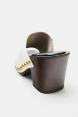 Zueco de piel color blanco, con tacón de madera y plataforma, detalle de tachas metálicas doradas en los laterales y punta cuadrada.