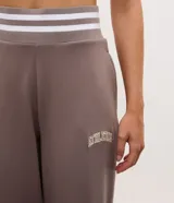 Pantalón deportivo de corte recto color marrón, confeccionado en una mezcla de modal y algodón. Presenta cintura elástica en rib con rayas blancas contrastantes, bolsillos laterales y detalle de lettering bordado en la pierna. Incluye aberturas laterales en el ruedo.