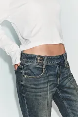 Jeans de tiro bajo con corte bootcut, confeccionados en denim con efecto lavado. Presentan costuras verticales marcadas en el frente, cierre frontal con cremallera, gancho metálico y detalle de trabilla con botones en la cintura.