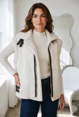 Chaleco estilo gamulán en color crudo, con interior y bordes de corderito sintético. Presenta cierre frontal con cremallera, detalles en cuero marrón oscuro y hebillas laterales ajustables.