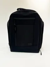 Mochila negra de forma rectangular, con múltiples cierres frontales y un pequeño parche rectangular con texto en relieve en la parte inferior central.