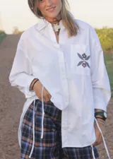 Camisa blanca de corte oversize y manga larga, con cuello clásico y cierre frontal de botones. Presenta un bolsillo en el pecho con un detalle bordado de flores en tonos marrones y azules, y cordones laterales decorativos.