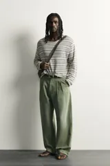 Pantalón chino baggy fit color verde militar, confeccionado en denim de algodón rígido, con corte holgado, tiro medio y pliegues frontales. Presenta bolsillos laterales y cierre frontal con cremallera y botón, con efecto lavado.