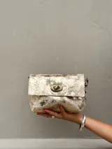 Cartera tipo bandolera de cuero con acabado metalizado en tono dorado envejecido. Presenta una solapa frontal con cierre giratorio metálico y herrajes en bronce. Incluye doble correa para usar al hombro o cruzada y un interior forrado con bolsillo.