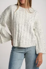 Sweater de punto color blanco roto con efecto tweed (pequeñas motas negras), cuello redondo y manga larga acampanada. Presenta un tejido texturado con trenzas verticales.
