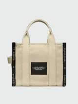 Bolso tipo tote Marc Jacobs modelo The Small Tote, color beige con detalles y texto en negro que dice "THE TOTE BAG MARC JACOBS". Cuenta con asas superiores y laterales para llevar en la mano o al hombro.