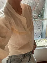 Camisa blanca a rayas verticales, con cuello redondo, mangas 3/4 abullonadas y detalle de puntilla en el pecho y mangas.