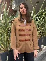 Chaqueta color tostado de estilo militar con detalles de tiras en cuero sintético marrón y botones metálicos decorativos en el frente. Presenta hombreras con detalle de tira y botón, mangas tres cuartos con puños vueltos y bolsillos frontales con solapa.