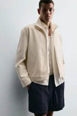 Cazadora de tejido color crema, con cuello alto, cierre frontal de cremallera y bolsillos de vivo en la cadera. Presenta un calce relajado (relaxed fit).