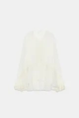 Blusa de escote en pico con manga larga terminada en volantes. Presenta aplicaciones de encaje a tono, cierre frontal con lazadas y bajo asimétrico.