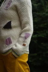Cardigan de lana merino tejido a mano con técnica de dos agujas, de color marfil con parches geométricos de tela en tonos celestes y lila aplicados con costuras visibles. Presenta cierre frontal con botones de madera y detalles de ribetes multicolores en los bordes.