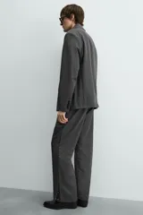 Pantalón de vestir gris oscuro, corte ancho (flare fit) y tiro alto, con bolsillos laterales y falsos bolsillos de vivo en la parte trasera. Presenta un cierre frontal con cremallera y gancho oculto, y botones a presión ocultos en el bajo.