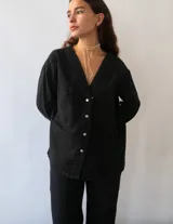 Camisa de denim negro con cuello en V, cierre frontal con botones metálicos y dos bolsillos cuadrados en el frente. Presenta un corte recto y oversize con ajuste en la espalda.