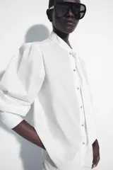 Camisa de algodón color blanco, con cuello subido y detalle de pañuelo incorporado, manga larga con puños anchos y cierre frontal con botones joya dorados.