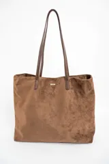 Bolso tipo tote de gran tamaño confeccionado en gamuza sintética color tostado. Presenta un diseño minimalista con asas largas de hombro y una pequeña placa metálica frontal con el logo de la marca.