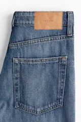 Jean de tiro alto confeccionado en denim de algodón rígido, con diseño de cinco bolsillos, perneras amplias y corte holgado. Presenta un lavado azul medio y detalle de dobladillo deshilachado.
