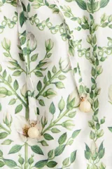 Vestido largo de algodón color blanco con estampado botánico en tonos verdes. Presenta cuello mao, cierre frontal con botones ocultos, mangas cortas abullonadas y falda de corte escalonado con volados. Incluye un cinturón fino para ajustar en la cintura.