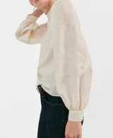 Blusa de color crema con cuello redondo, mangas largas abullonadas y detalles de bordado y puntilla vertical en el frente.