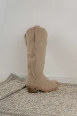 Bota estilo tejana de caña alta, confeccionada en gamuza, con taco cuadrado y diseño de costuras decorativas.