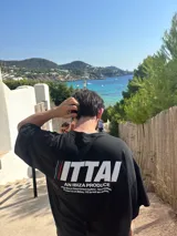 Remera marrón de corte holgado con estampado en la espalda que incluye la palabra "ITTAI" en blanco con líneas rojas y azules, la frase "AN IBIZA PRODUCE" y un texto más pequeño que dice "Ittai Stars Department Gallery - Ibiza Edition. If you're in Ibiza, 112 is not an option.".