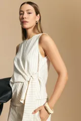 Top sin mangas color beige con finas rayas verticales negras. Presenta un diseño con lazada ajustable en el lateral de la cintura.