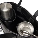 Cartera tipo tote bag de cuero sintético negro con textura granulada. Cuenta con doble asa superior, correa ajustable extraíble, bolsillo frontal con cierre metálico y logo de la marca grabado en la parte inferior.