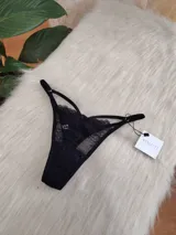 Tanga de encaje negro con diseño de tiras laterales regulables y detalle de transparencia. Cuenta con refuerzo de algodón en la entrepierna.