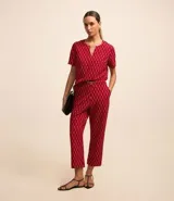 Blusa de crepé color rojo con estampado geométrico de rombos en negro y blanco. Tiene cuello redondo con abertura en forma de gota y detalle metálico, mangas cortas y corte recto.