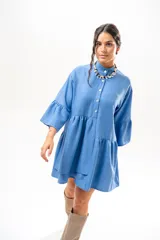 Vestido corto azul, estilo camisero, con cuello camisero, cierre frontal con botones y mangas acampanadas con volados. Presenta un diseño holgado con cortes fruncidos en el cuerpo.