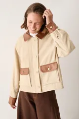 Campera corta en tono beige con cuello, puños, solapas de bolsillos frontales y detalles en cuero sintético marrón. Cuenta con cierre frontal de botones dorados y bolsillos aplicados.