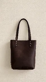Cartera tipo tote bag de cuero marrón, con diseño minimalista, asas largas y detalles de remaches metálicos en la unión de las asas.