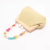 Cartera de mano con estructura rígida de madera clara, cuerpo de tejido de paja trenzado color verde claro y cierre de dos esferas blancas. Incluye un asa desmontable compuesta por cuentas irregulares de resina en tonos pastel y vibrantes.