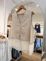 Chaleco beige con escote en V, un botón frontal y un detalle de bajo en gasa blanca que crea un efecto de capas.