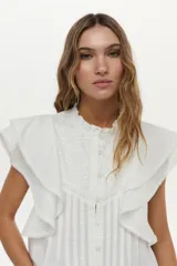 Blusa blanca sin mangas, con volados amplios en los hombros, bordado a tono en el pecho, detalles de tablones a lo largo del cuerpo y cuello elevado con volados.